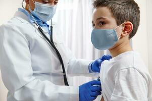 Doctor pediatrician in medical uniform uses a stethoscope while auscultating a little boy in protective medical mask photo