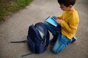 Charming elementary aged schoolboy sits on his knees on a path in the park, puts a notebook and pencil case in a backpack, returning home after shooting photo