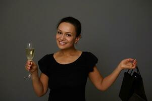 Beautiful African woman shopper smiles looking at camera posing against gray background with a champagne flute and shopping black packet. Concept of celebration the Black Friday with copy space for ad photo