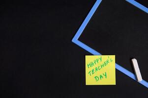 Flat lay of a pasted yellow note paper with inscription Happy Teacher's Day on an empty blank board with a chalk. Black background, copy space photo