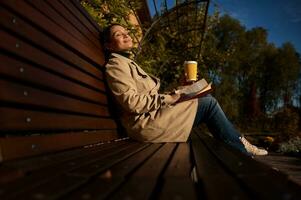 Attractive woman with closed eyes sits on wooden bench in park, holding paper cup of hot drink and a book, enjoys autumn sunbeams, away from the hustle and bustle from the city, resting from gadgets photo