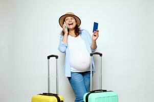 Happy pregnant woman talking on mobile phone, booking hotel, posing with boarding pass and suitcases on white background photo