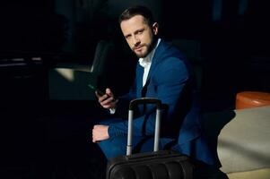 Portrait of successful confident happy young businessman looking at camera sitting on armchair with smartphone in hotel lobby. Business trip concept photo