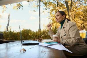 Pensive pretty business lady holds a pen in one hand and takeaway paper coffee cup in the other and thoughtfully looks the side while working remotely on laptop in the outdoor cafe at cool autumn day photo