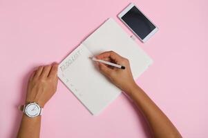 High angle view of female hands holding pencil, writing in notebook, checking to-do list. Mobile phone lying on pink background with space for text photo