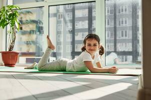 adorable niña relajante en un ejercicio estera y sonrisas mientras posando a cámara en contra el antecedentes de grande ventanas a hogar en un soleado día foto