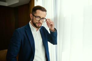 Waist-length portrait of a successful business investor, handsome successful young businessman standing by the window in a hotel room and looking confidently at the camera through eyeglasses photo