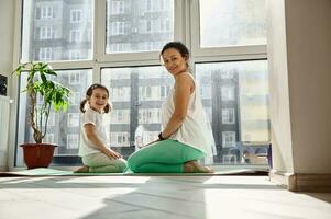 Charming young mother and her daughter sitting on a yoga mat against the background of large windows and smiling to camera. photo