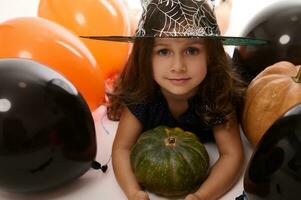 Close-up portrait of pretty little girl in carnival witch costume and wizard hat looking at camera posing with pumpkin in the white background with colored black and orange balloons. Halloween concept photo