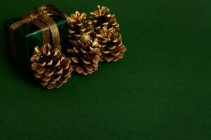 de cerca composición para nuevo año y Navidad anuncio de dorado pino conos y pequeño lujo presente en Brillantina verde envase regalo papel con dorado arco en el esquina de verde antecedentes con Copiar espacio foto