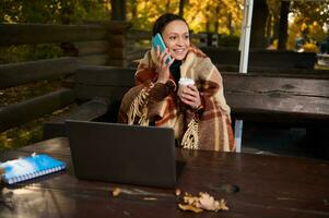 Beautiful woman, developer wrapped in a warm plaid woolen blanket, talks on mobile phone, holds a takeaway paper cup with hot coffee while working remotely on laptop, planning projects, start up photo
