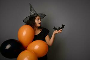 Beautiful African American woman in wizard hat, dressed in black, holds orange balloons and a handmade felt-cut black bat, poses against gray background with copy space for ad. Halloween party concept photo