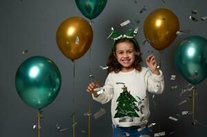 Cheerful baby girl in elf hoop having fun at New year party throwing sequins confetti, rejoicing against gray background with shiny golden green inflated air balloons. Christmas concept, copy ad space photo