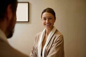 Beautiful middle aged European woman in bathrobe smiling looking at her blurred husband on the foreground, resting at wellness spa resort on honeymoon. Cute couple getting beauty treatment together photo