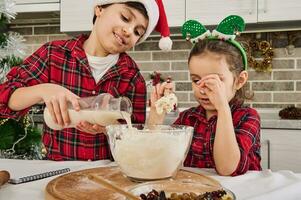 Handsome preadolescent Caucasian boy dressed in red and green checkered shirt and Santa hat pours some milk into a bowl, cooking with his little sister Christmas cake at home kitchen. photo