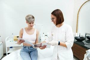 simpático cosmetóloga demostración un catalogar de precios para cosmético procedimientos a un rubia cliente sentado en un Sillón en un belleza salón durante un consulta a un belleza spa clínica foto