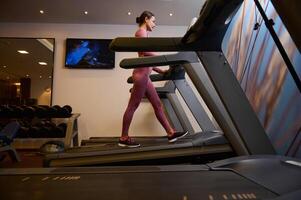 Side view of a middle aged European athlete woman with perfect physique in sportswear exercising on a treadmill at gym. Cardio workout. photo