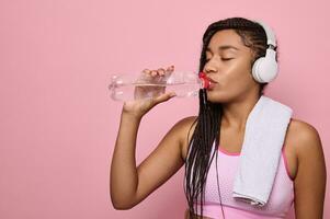 Fitness woman drinking water standing on pink background with copy space. Portrait of African cute woman taking a break after intense workout. Mid adult lady drink from water bottle after gym workout photo