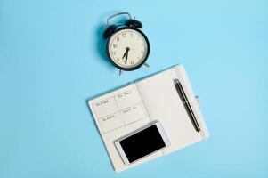 Flat lay of a mobile phone smartphone and an ink pen on an open notebook with schedule next to a black alarm clock, isolated on color background with copy space. Time management and schedule concepts photo