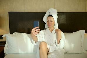 Happy young Caucasian woman in white bathrobe and wrapped head in bath towel lying on the bed in hotel bedchamber and smiles looking at camera on her smartphone while taking self-portrait, selfie photo