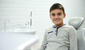 adorable niño preadolescente chico sentado en del dentista silla durante dental cita para regular médico preventivo chequeo, sonrisas con dientes sonrisa mirando a cámara. dental y oral cuidado concepto foto