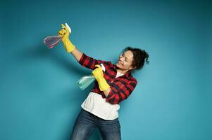 Young woman playing with cleaning sprays as if shooting a pistol. Posing over blue background with copy space. Cleaning and home work concept photo
