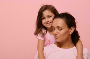 hermosa bebé niña en rosado vestir con un rosado cinta tiernamente abrazos su madre con cerrado ojos, mirando a cámara, demostración apoyo a cáncer pacientes y supervivientes, rosado antecedentes con Copiar espacio foto