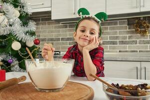 Adorable child, beautiful baby girl smiles, dreamily looking at the camera, mixing dry ingredients in a glass bowl. Cute little chef prepares cookies and sweets for christmas traditional events photo