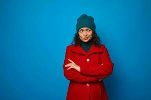 Portrait of a puzzled thoughtful pensive curly dark-haired pretty African American woman in red coat and green looking aside posing with crossed arms against blue wall background with copy ad space photo