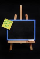 Close-up of a wooden table easel with chalk and blackboard with empty blank space for text and pasted note paper with inscription teachers day isolated on black background with copy space photo