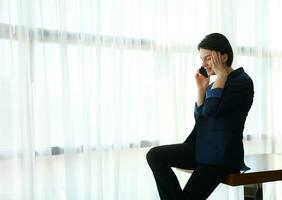 Young brunette woman, CFO, manager at business trip, wearing business casual suit talks on mobile phone sitting on a table on the background of large hotel windows photo