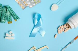 Flat lay of Blue Diabetes Awareness ribbon in center of blue background with scattered pharmaceutical pills, insulin syringe, caliper, measuring tape, blister with medicines and refined white sugar photo