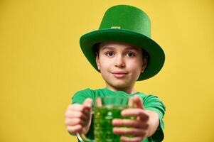 A boy in leprechaun hat holds a glass of green drink out in front of him to the camera. Isolated on yellow background. Saint Patrick day photo