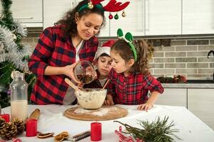 hermosa contento amoroso madre y su adorable alegre niños, hijo y hija Cocinando juntos un Navidad pastel masa en el cocina. felicidad es a cocinar con mamá foto