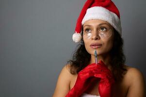 Beautiful woman with natural make-up and patches under eyes, in Santa hat holds syringe with beauty injection near her face, looks up on copy space of gray background. Injection cosmetology concept photo