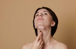 Young brunette woman doing a lifting massage on her neck, using a jade roller massager. Beauty portrait on beige background with space for ad. photo