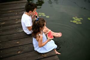 Kids sit on the pier, lowered their feet in the pond with water lilies, relax in nature, in the fresh air, eat fresh ripe watermelon photo