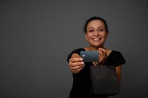 Soft focus on black credit card in the hand of smiling joyful woman dressed in black and posing with black shopping packet against gray wall background with copy space for Black Friday ad photo