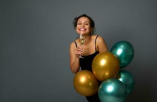 Attractive African American woman smiles looking at camera, poses with champagne flute and beautiful festive shiny golden and green metallic air balloons against gray wall background with copy space photo