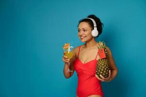 Smiling mixed race brunette in swimsuit holding pineapple with blank red card and cocktail, enjoying music with headphones photo