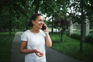 Beautiful multi-ethnic young woman talking on smartphone while strolling the alley of a city park on a summer day photo