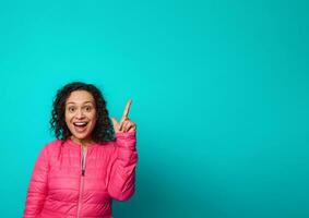 Surprised cheerful brunette in bright pink jacket looks at camera and points with her finger up to a copy space for advertisement text on blue background. photo