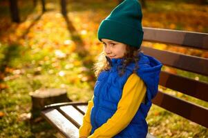 Encantado maravilloso bebé niña en vistoso calentar ropa y azul acogedor chaqueta sentado en de madera banco, lindo sonrisas en el antecedentes de rayos de sol que cae en otoño parque con caído seco hojas a puesta de sol foto