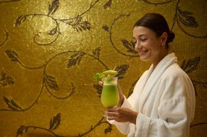 Smiling middle aged Caucasian pretty woman in white bathrobe, holding a glass with delicious healthy freshly squeezed vitamin juice in her hands, standing in a luxurious wellness center lounge area photo