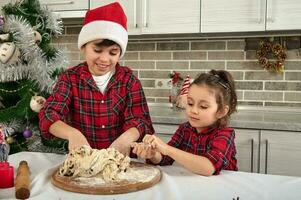 hermosa niños Cocinando juntos en el hogar cocina durante Navidad vacaciones. adorable chico en Papa Noel sombrero y su linda pequeño hermana preparando y amasadura masa para un pan foto