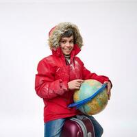Adorable school boy in a warm snow-covered red parka with a hood, sitting on a suitcase, smiles toothy smile pointing his finger at the globe in search of a travel destination. Winter tourism concept photo