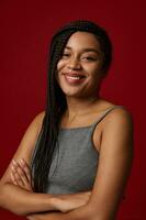 Young adorable African american woman with long pigtails dreadlocks in gray sports t-shirt smiling cute toothy smile while looking at camera, posing against red colored background with copy space photo