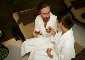 Handsome attractive man and beautiful gorgeous woman, both of them wearing white cozy terry bathrobes, resting in lounge zone by pool in luxury wellness spa centre, cutely talking over cup of tea photo