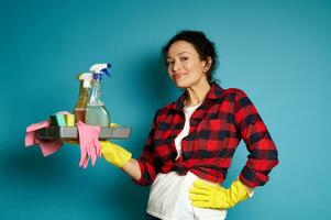 A curly brunette wearing a white T-shirt, a red check shirt and yellow rubber gloves, holds a tray of cleaning supplies and cute smiles while posing on a blue background with copy space photo