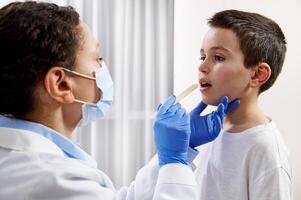 African American doctor with face mask and protective gloves examining caucasian boy's throat during a home visit. photo